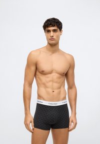 Mies, joka ei ole paidassa ja jolla on lyhyet kiharat hiukset, pukeutuneena mustiin kuviollisiin Calvin Klein -boxerihousuihin, seisomassa vaaleataustaista taustaa vasten.