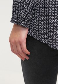 Svart skjorta med vitt triangelmönster, knäppte manschetter och mörkgrått tyg. Visar en hand som vilar mot plaggets kant.