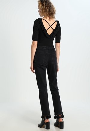 Femme debout de dos portant un haut noir avec un dos en V volanté et des bretelles croisées, un jean noir avec des pampilles aux poignets et des chaussures à talons noires.