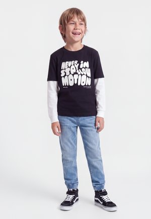 Smiling boy wearing a black t-shirt with white text "Never stay in slow motion," light blue jeans, black and white sneakers, standing on white background.