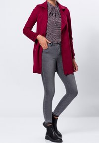 Bordeaux suede jas over een gepatterde blouse, gecombineerd met grijze skinny jeans en zwarte enkellaarsjes. Gladde textuur met een getailleerde pasvorm.