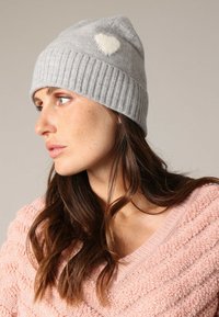 Grijze gebreide beanie met ribbelstructuur en witte hartdetail aan de zijkant. Zacht materiaal, aansluitend ontwerp en afgeronde bovenkant.