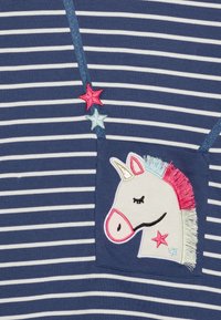 Námořnická a bílá pruhovaná látka obsahuje nášivku s motivem jednorožce. Jednorožec má růžové a modré detaily a nad ním jsou vyšívané hvězdy.