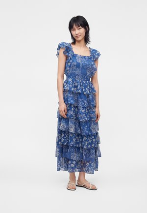 Femme debout dans une robe bleu fleurie à étages avec manches à volants et sandales plates blanches à brides sur fond blanc.