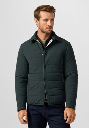 Man wearing dark green quilted jacket with snap buttons over layered shirts and dark jeans, standing against plain background.