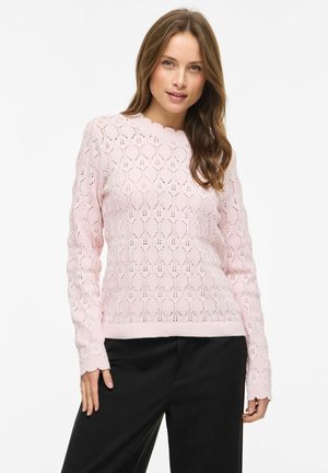 Kvinde med langt brunt hår iført en lyserød, langærmet strikket sweater med bølget halsudskæring og sorte bukser, stående mod en hvid baggrund.