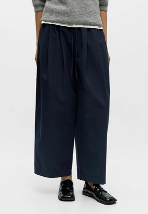 Person trägt weite marineblaue Hosen, grauen Pullover, weißes Unterhemd und schwarze glänzende Loafers und steht vor weißem Hintergrund.