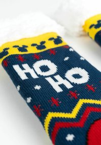 Calze a maglia con una base blu navy, testo bianco "HO HO", dettagli gialli e rossi, e un morbido polsino in pelliccia bianca. I motivi includono fiocchi di neve rossi.