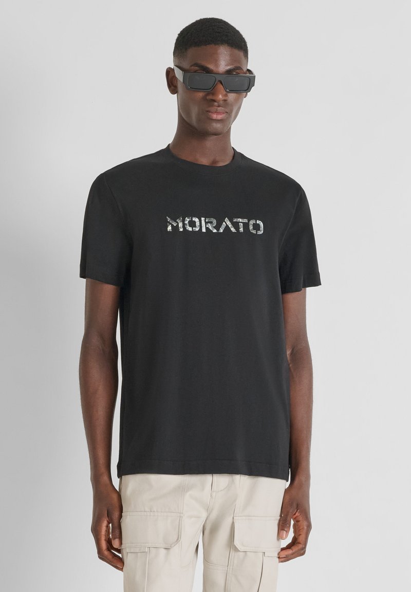 Camiseta de algodón negra con un logo reflectante "MORATO" en el pecho. Diseño de mangas cortas y cuello redondo, combinada con pantalones cargo beige.