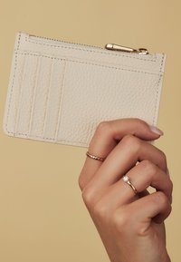 Cream leather cardholder with four slot sections and a top zipper. Features textured surface and gold-toned hardware accents.