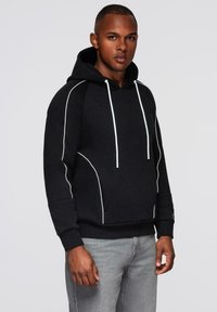 Junger Mann in einem schwarzen Hoodie mit weißen Abnähern und Kordeln, kombiniert mit hellgrauen Jeans, steht vor einem schlichten Hintergrund.