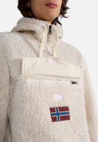Pullover in pile color crema con colletto alto, presenta una tasca frontale con pattina, zip nera e una toppa con la bandiera norvegese.