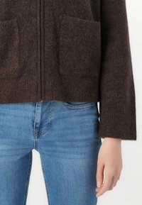Personne portant un pull zippé en velours brun avec deux poches avant et un jean en denim bleu clair, la main reposant sur le côté.