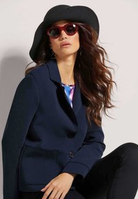 Marineblauen Blazer mit Reverskragen, einem Knopf und aufgesetzten Taschen, kombiniert mit einem Blumenoberteil. Accessoires sind ein schwarzer Filzhut und rote Sonnenbrille.