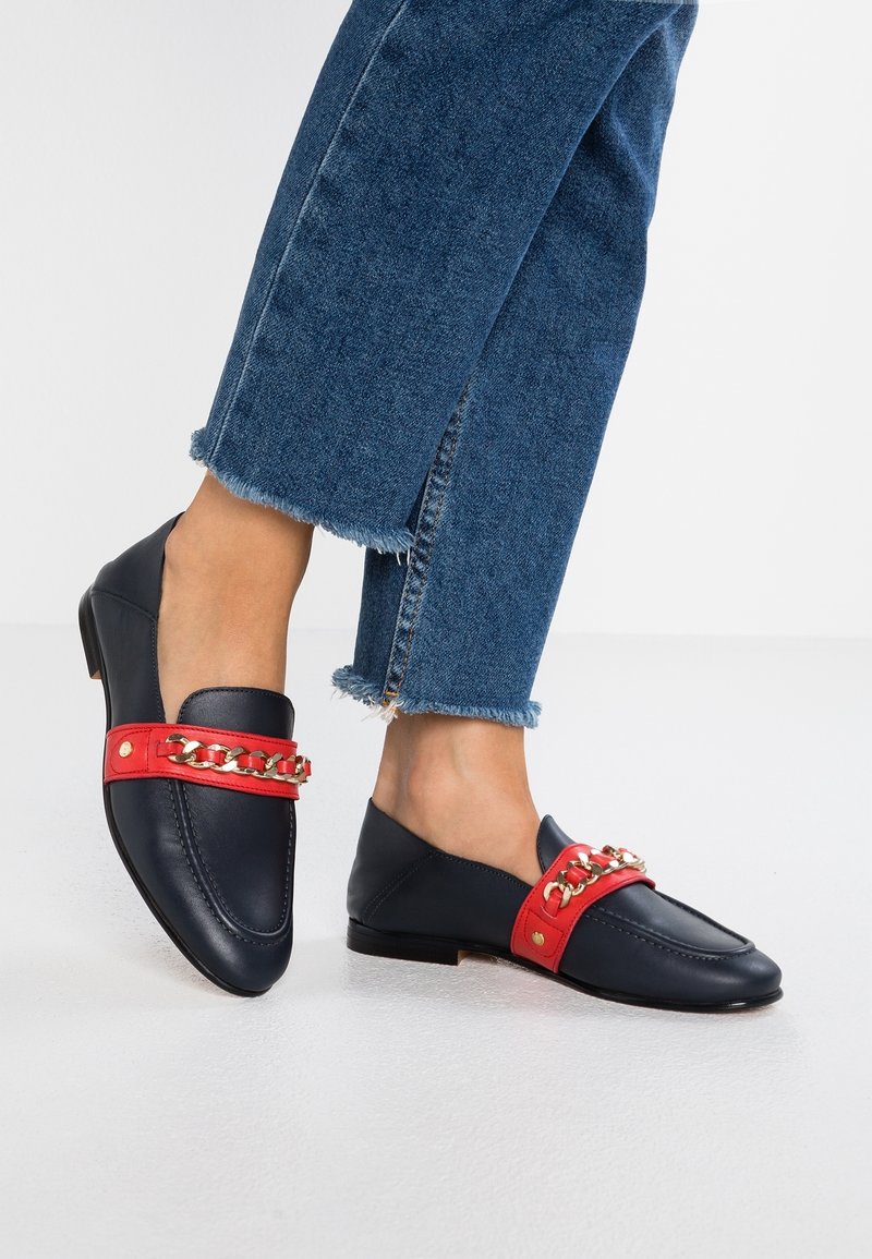 Mocassins en cuir bleu marine avec une bride rouge ornée d'accents en chaîne dorée, accompagnés d'un jean en denim à ourlets effilochés.