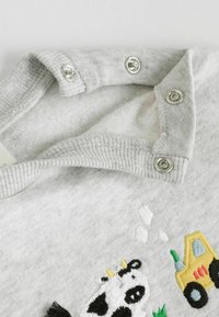 Hellgraues Babybekleidungsstück mit Druckknöpfen und gesticktem Patch eines Cows sowie einem gelben Traktor mit grünem Gras.