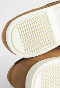 Brązowe skórzane sneakersy z białymi gumowymi podeszwami, wyposażone w teksturowany wzór w zygzak. Cholewka ma widoczne przeszycia i gładkie wykończenie.