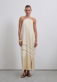 Robe maxi en soie crème avec des bretelles spaghetti, des détails en franges et une fente latérale. Portée avec des sandales plates noires et un bracelet jaune.