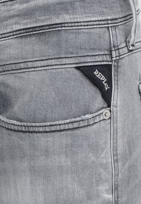Grå denimjeans med en bakficka som har en marinblå etikett märkt "REPLAY" och en silverfärgad knappdetalj. Slät textur med urblekta områden.