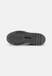 Grå sneakersål med tekstureret gummioverflade, der har et zigzag-mønster, riller for bedre greb og en central udskæring for fleksibilitet.