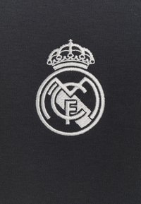 Logo de Real Madrid bordado en blanco con una corona sobre un fondo de tela oscura. El diseño incluye elementos circulares y geométricos.