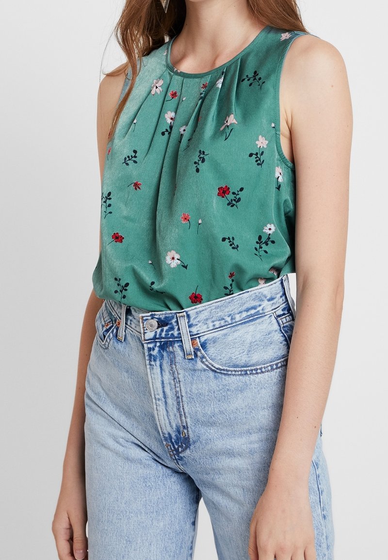 Top senza maniche verde a fiori con plissettature, caratterizzato da piccoli fiori rossi e bianchi, abbinato a jeans in denim blu chiaro a vita alta.