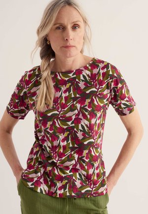 Femme avec une longue tresse blonde portant un haut à manches courtes fleuri avec des fleurs roses et des feuilles vertes, associée à un pantalon vert, debout les mains dans les poches.