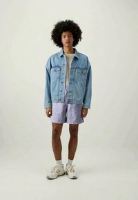 Light blue denim jacket with two chest pockets, white t-shirt, lavender patterned shorts, and beige athletic shoes. Short, curly hair.