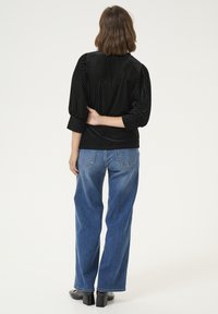 Camicia di velluto nero con texture a coste verticali, maniche ¾, abbinata a jeans blu a vestibilità comoda. Il modello sta in piedi con le braccia incrociate in vita.