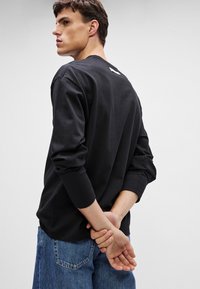 Svart langermet T-skjorte i bomull med rund hals, med et subtilt logomerke bak, kombinert med lyseblå jeans.