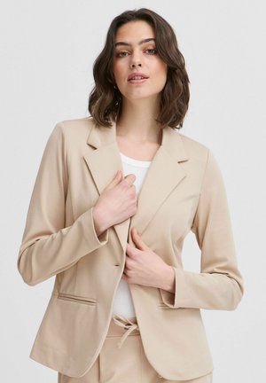 Beige přizpůsobený blazer s klopami, jednobotovým zapínáním, předními kapsami a hladkým materiálem. Nosený přes bílý top s rovnými rukávy.