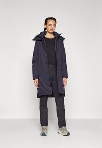 Cappotto lungo blu navy con cappuccio, dotato di chiusura con zip e bottoni decorativi, abbinato a pantaloni scuri e sneakers spesse di un colore neutro.