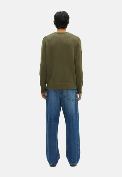Hombre de pie de espaldas, vistiendo un suéter verde oliva desgastado y jeans holgados de color azul, contra un fondo claro y liso.