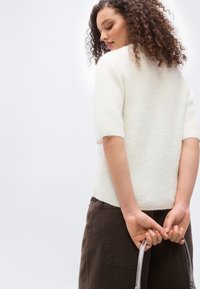 Γυναίκα με σγουρά μαλλιά, φοράει λευκό πουλόβερ με κοντά μανίκια και σκούρο παντελόνι, κρατώντας μια γκρι λαβή πίσω από την πλάτη της, κοιτάζοντας προς τα κάτω.