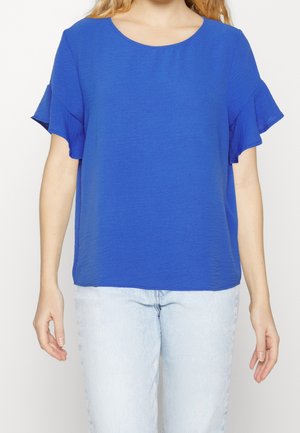 Femme portant une blouse bleu roi à manches courtes avec des détails de volants, associée à un jean en denim clair.