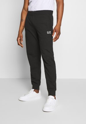 Homme portant un pantalon de jogging noir EA7 Emporio Armani et des baskets blanches, debout devant un fond gris uni.