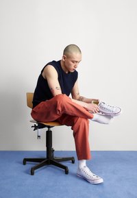 Maglione nero senza maniche, pantaloni arancioni, sneaker bianche con accenti rossi. Seduto su una sedia girevole in legno contro uno sfondo semplice.