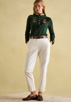 Grünes Langarm-Polo mit goldenen Akzenten, Seitenpaneel-Design und bestickten Details. Kombiniert mit weißen Hosen und braunen Leder-Mokassins.