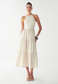 Beige halterneck-mekko, jossa on kerroksellinen hame, kevyt kangas, pehmeä texture ja epätasainen helma. Yhdistetty valkoisten nyörisaappaiden ja rannekorun kanssa.