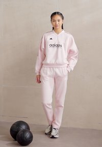 Roze sportoutfit bestaande uit een sweatshirt met ronde hals en zwart logo, met bijpassende joggingsbroek. De outfit wordt aangevuld met grijze sportschoenen.