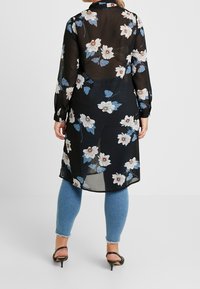 Gennemsigtig sort lang skjorte med et blomsterprint i hvidt og blåt, parret med lyseblå jeans og sorte hælesandaler.