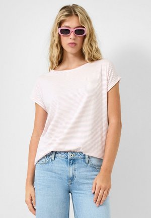 Kobieta o długich blond włosach, nosząca różowe okulary przeciwsłoneczne, różowy jasnoróżowy top w paski z krótkim rękawem oraz jasnoniebieskie dżinsy, stojąca na jednolitym tle.