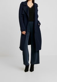 Granatowy płaszcz trencz o dwurzędowym kroju, z regulowanymi mankietami i bocznymi kieszeniami, noszony na czarnej bluzce z koronkowym wykończeniem i szerokich spodniach w kolorze morskim.