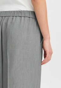 Grijze broek met een zachte textuur, voorzien van een geplooide tailleband en zijzakken, weergegeven met een hand die rust nabij de naad.