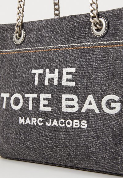 Sac fourre-tout en denim gris avec des anses en chaîne argentée. Il présente un texte blanc indiquant "THE TOTE BAG" et "MARC JACOBS" imprimé sur le devant.