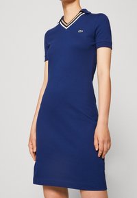 Marineblaue Polokleid mit kurzen Ärmeln, das einen V-Ausschnitt mit beige-weißen Streifen und ein kleines grünes Logo auf der Brust hat.