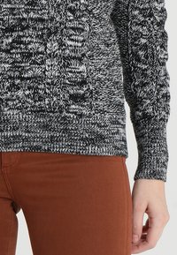 Strickpullover in Schwarz und Weiß mit strukturiertem Muster und gerippten Bündchen, kombiniert mit rostfarbenen Hosen.