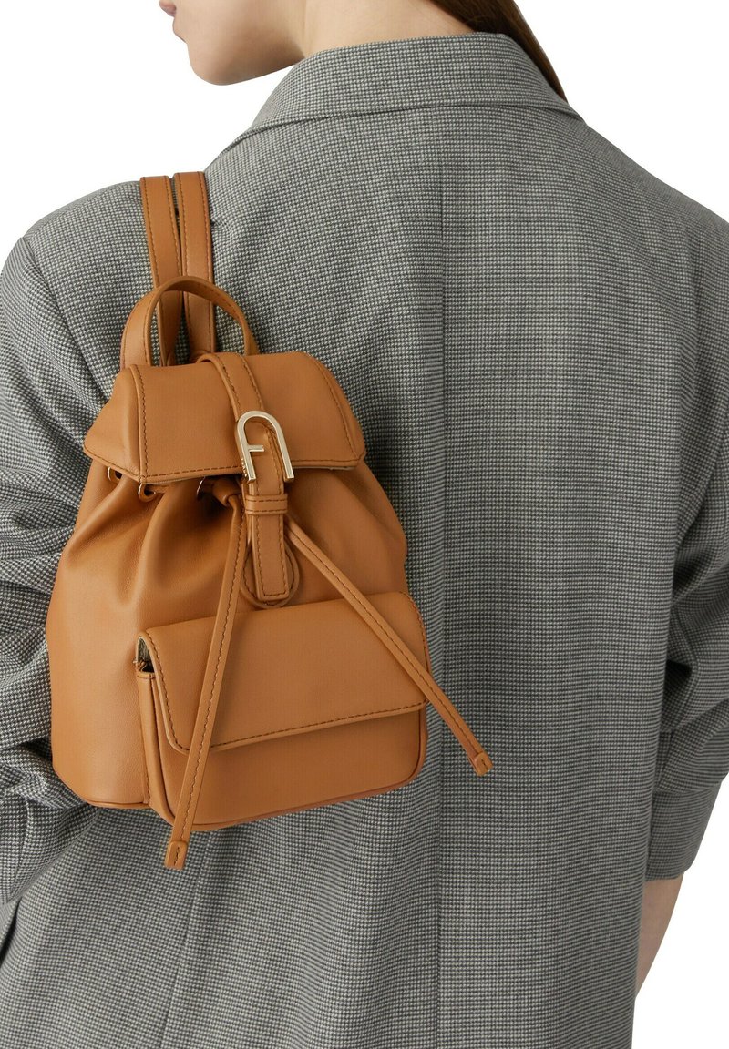 Mochila de cuero marrón con solapa superior, correas ajustables y un bolsillo delantero, con un cierre de hardware dorado. Llevada sobre una prenda de cuadros gris.