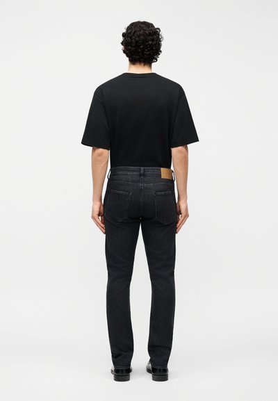 Άνδρας που στέκεται με την πλάτη γυρισμένη, φορώντας ένα απλό μαύρο μπλουζάκι με κοντά μανίκια, σκούρα τζιν με πίσω τσέπες και μαύρα παπούτσια μπροστά σε απλό φόντο.