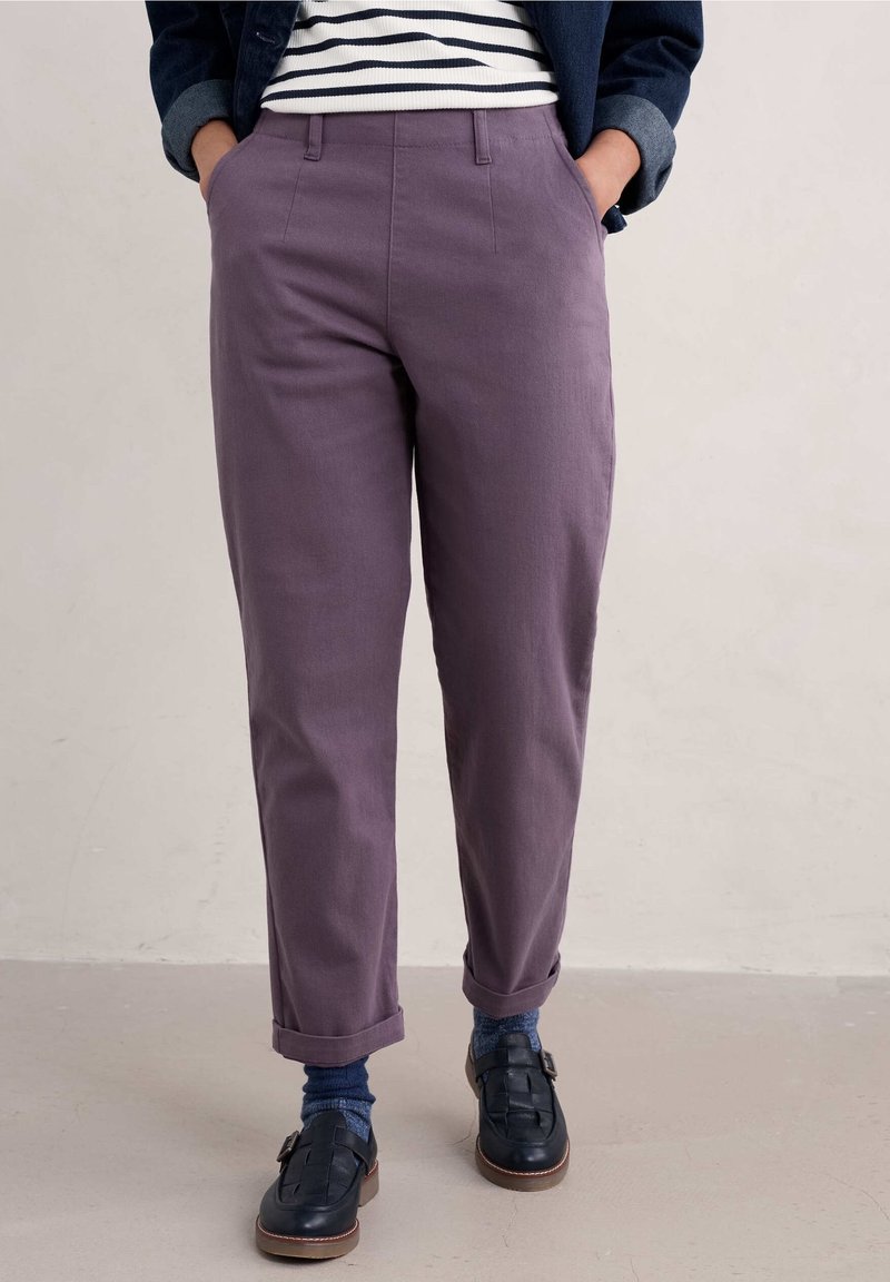 Lila raka byxor med framfickor, som har en slät tygtextur. Uppvikta vid anklarna, kombinerade med mörka skor och blå strumpor.